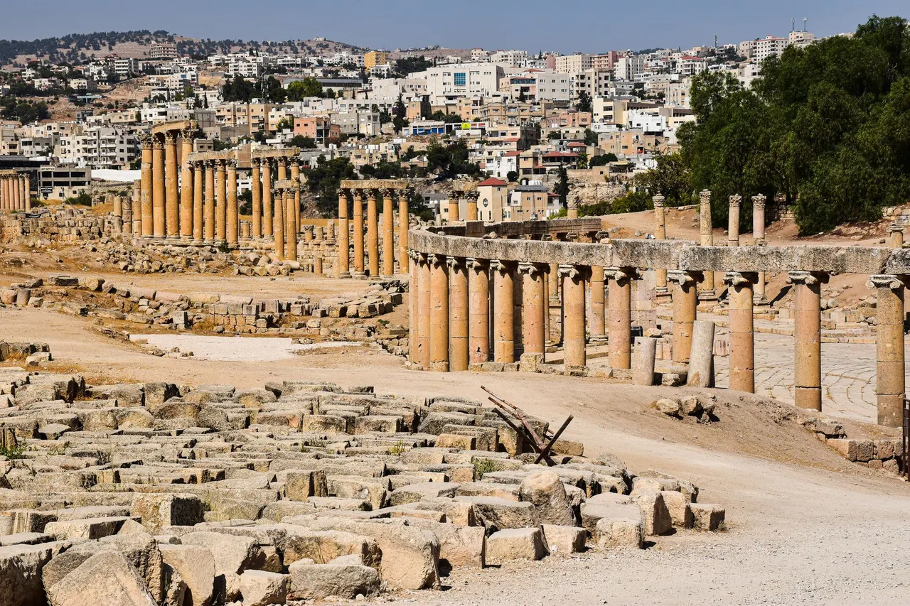 الساحة البيضاوية في جرش: صفّ أعمدتها الأيونية المنحني في المقدّمة، وأعمدة الشارع المعمَّد خلفها، ومدينة جرش الحديثة على التلّة فوق الموقع